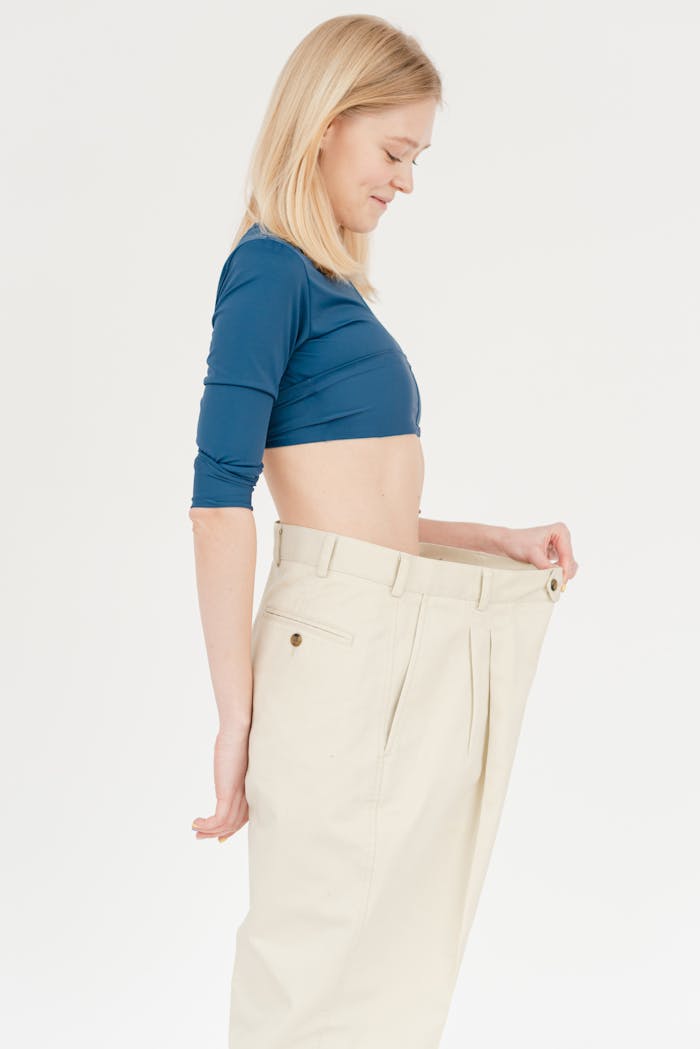 Side view of slim positive female with bare belly wearing oversized trousers standing on white background in light studio after weight loss